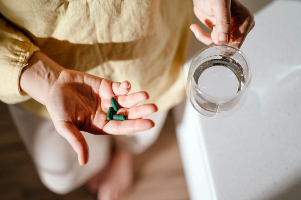 A person is holding green capsules in one hand and a glass of water in the other, perhaps preparing to take supplements or medicine.