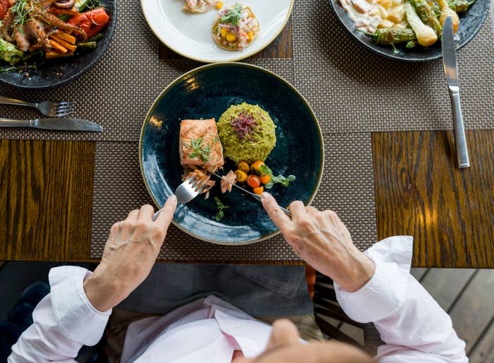 A person eating a dish of salmon, a piece of green rice, and mixed vegetables, surrounded by other dishes on the table.